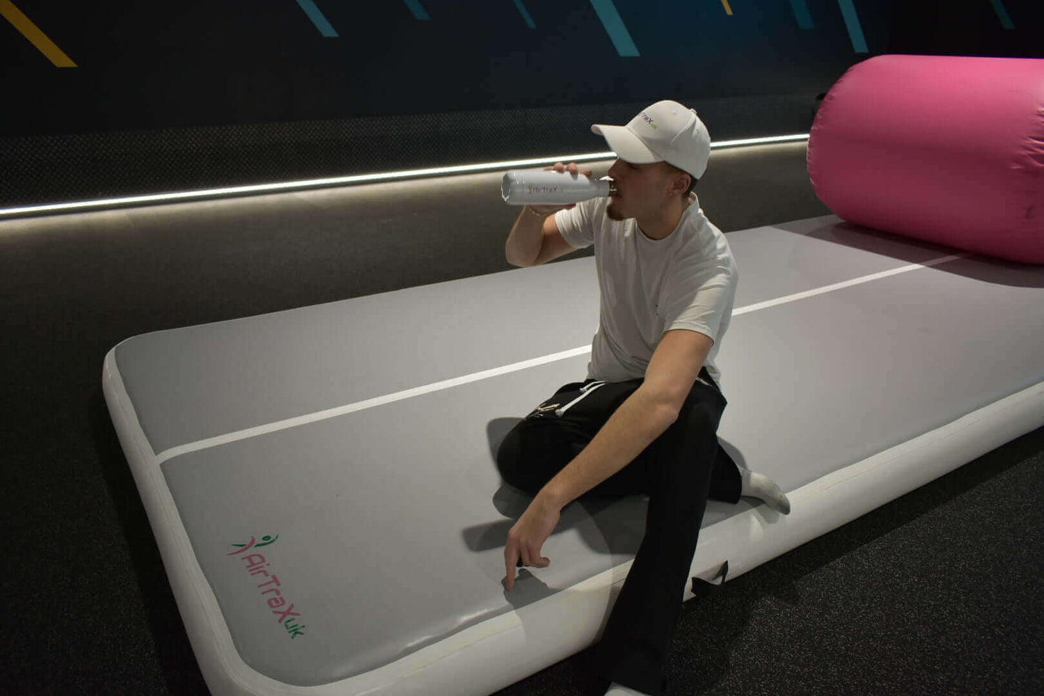 Person sitting on AirTraX Pro air track mat, drinking water in a fitness setting.